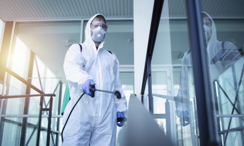 Person in white chemical protection suit doing disinfection of public areas to stop spreading highly contagious corona virus.